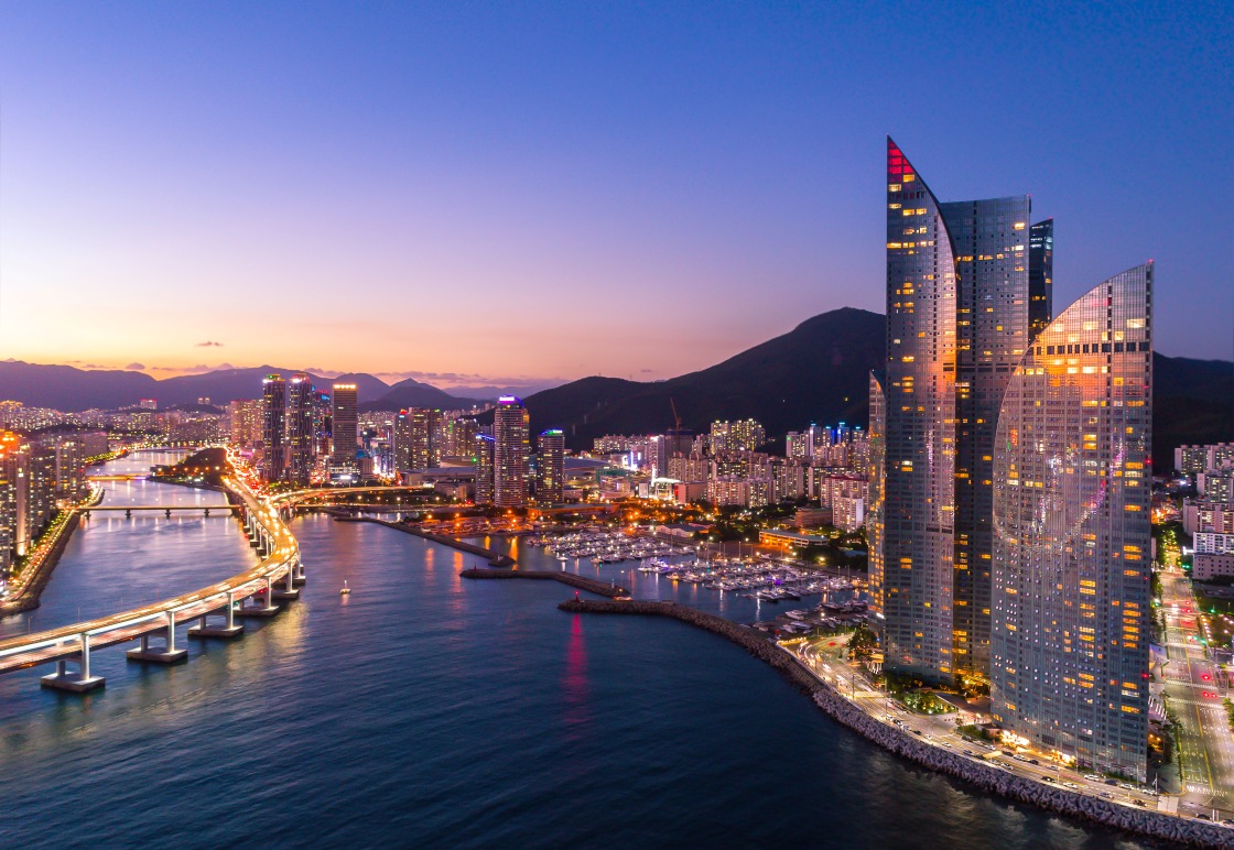 Marine City glows at dusk as Gwangan Bridge illuminates the harbor and Park Hyatt Busan rises along the right — a sculptural glass landmark mirroring the arc of the waterfront. © Chai Photographer/Shutterstock