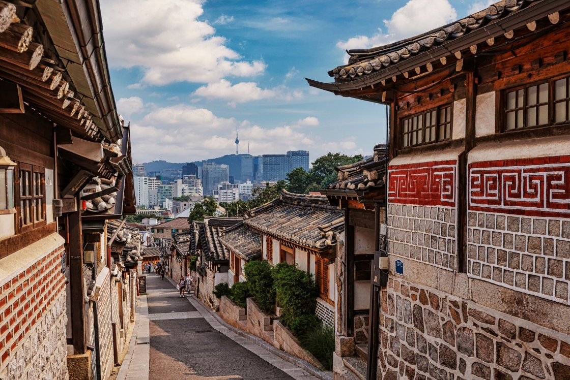 Traditional hanok line the narrow lanes of Bukchon Hanok Village, with Seoul’s modern skyline rising in the distance. © kdream/Shutterstock