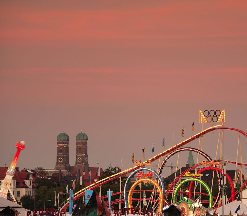 Das Oktoberfest in der Dämmerung mit dem Wahrzeichen der Stadt München, der Frauenkirche, im Hintergrund sowie dem Klassiker der Wiesn, dem Olympia-Looping © München Tourismus, B. Roemmelt